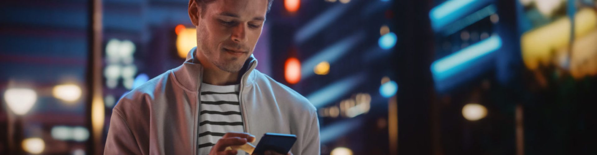 Handsome Man Using Smartphone Walking Through Night City Full of Neon Colors and Entertainment. Stylish Young Man Using Mobile Phone, Posting on Social Media, Online Shopping, Texting on Dating App